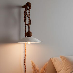 May include: A hanging pendant light with a white, textured shade and a warm, glowing bulb. The light is suspended by a brown rope and wooden accents, creating a rustic and natural aesthetic. The design is suitable for a bedroom or living room.