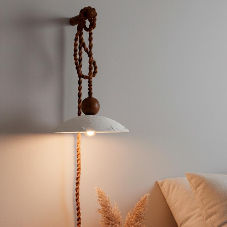 May include: A hanging pendant light with a white, textured shade and a warm, glowing bulb. The light is suspended by a brown rope and wooden accents, creating a rustic and natural aesthetic. The design is suitable for a bedroom or living room.