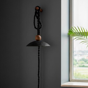 May include: A black pendant light fixture with a textured, rounded shade and a wooden accent. The light hangs from a black rope and is mounted on a wooden peg. The fixture is against a dark gray wall.