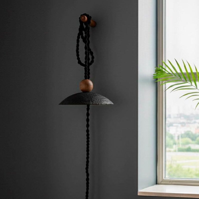 May include: A black pendant light fixture with a textured, rounded shade and a wooden accent. The light hangs from a black rope and is mounted on a wooden peg. The fixture is against a dark gray wall.