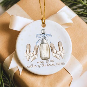 Pode incluir: Enfeite de Natal redondo em cerâmica branca com fio dourado. O enfeite apresenta uma garrafa de champanhe com um laço azul e três pares de saltos altos. O texto diz "mãe EST 1995 mãe da noiva EST 2025".