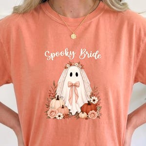 May include: A coral-colored t-shirt featuring a graphic of a ghost wearing a bridal veil and a bow. The text Spooky Bride is printed above the ghost, which is surrounded by pumpkins and flowers in shades of orange, pink, and white.