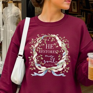 Puede incluir: Una sudadera de cuello redondo color granate con un gr&aacute;fico central de una corona floral hecha de hojas verdes y cintas azul claro. El texto dentro de la corona dice HE RESTORES my soul PSALM 23:3 en fuentes serif y manuscrita.