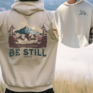 Op de afbeelding: Een beige hoodie met een grote achteropdruk van een berglandschap met bomen, een meer en vogels, met de tekst "BE STILL" en "PSALM 46:10" eronder. Op de borst staat een kleinere afbeelding van een berg en een kruis.