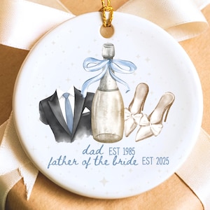 May include: A round, white ceramic ornament with a gold hanging string. The ornament has a watercolour design of a tuxedo, champagne bottle with a blue bow, and high heels. Text reads "dad EST 1985 father of the bride EST 2025."