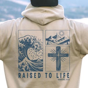 Op de afbeelding: Beige hoodie met een blauw grafisch ontwerp op de rug. Het ontwerp bevat een golf, bergen en een kruis, met de tekst "RAISED TO LIFE" en "ROMANS 6:4". De hoodie heeft een capuchon en een trekkoord.