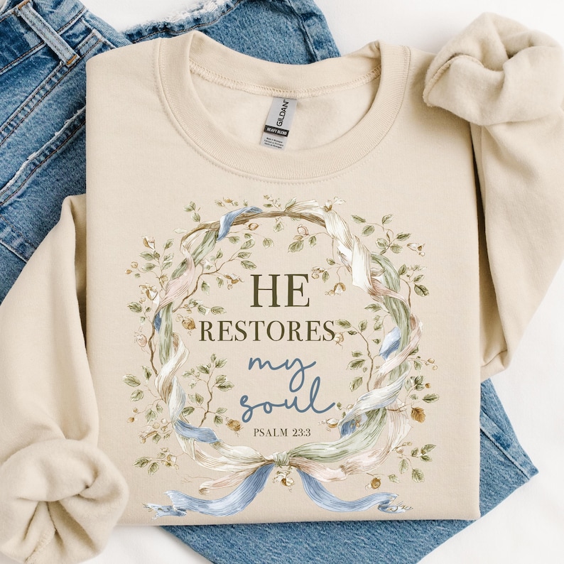 Puede incluir: Una sudadera de cuello redondo color arena con un gr&aacute;fico central de una corona floral entrelazada con cintas azul claro y blancas. El texto dice HE RESTORES my soul PSALM 23:3 en una combinaci&oacute;n de fuente serif gris oscuro y caligraf&iacute;a azul.