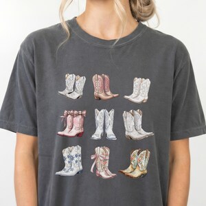 May include: Dark grey t-shirt with a watercolour illustration of nine cowboy boots. The boots are in various colours, including white, pink, and brown, with floral and decorative designs. The shirt is a casual, comfortable style.