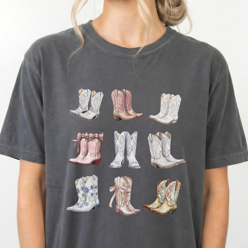 May include: Dark grey t-shirt with a watercolour illustration of nine cowboy boots. The boots are in various colours, including white, pink, and brown, with floral and decorative designs. The shirt is a casual, comfortable style.