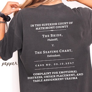 Puede incluir: Una camiseta de manga corta gris carbón con texto blanco en la espalda: IN THE SUPERIOR COURT OF MATRIMONY COUNTY, THE BRIDE, Plaintiff, V. THE SEATING CHART, Defendant. CASE NO. 09.10.2027, COMPLAINT FOR EMOTIONAL DISTRESS, UNFAIR PLACEMENT.