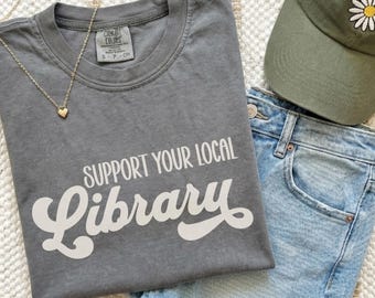 以下が含まれることがあります： 前面に白い文字で「SUPPORT YOUR LOCAL Library」とプリントされたチャコールグレーの半袖Tシャツ。クルーネックで「Comfort Colors」のタグが付いています。ゴールドトーンのチェーンネックレス、ライトブルーのデニムショートパンツ、白いデイジーの刺繍が施されたオリーブグリーンのベースボールキャップも写っています。