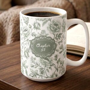以下が含まれることがあります： 緑と白の花柄が施された、濃いコーヒーが入った白いセラミックマグ。マグには大きなハンドルがあり、楕円の中に「Chapter 50」の文字が入っています。マグは木製の表面に置かれています。