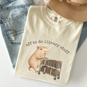 May include: Cream t-shirt featuring the text "off to do library stuff" and a capybara graphic pushing a book cart. The shirt is styled with light blue jeans and a beige hat. The shirt is made of a soft material.