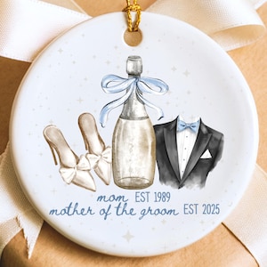 May include: A round, white ceramic ornament with a gold string for hanging. The ornament features a champagne bottle with a blue bow, a pair of high heels, and a tuxedo. Text reads "mom EST 1989" and "mother of the groom EST 2025".
