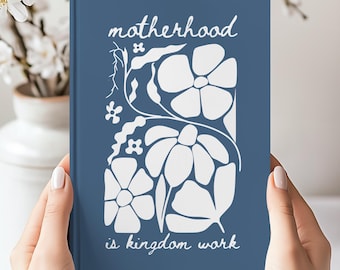 May include: A blue hardback journal featuring a white graphic print of stylised flowers and vines. The text "motherhood" is printed in white at the top, and the text "is kingdom work" is printed in white at the bottom of the front cover.