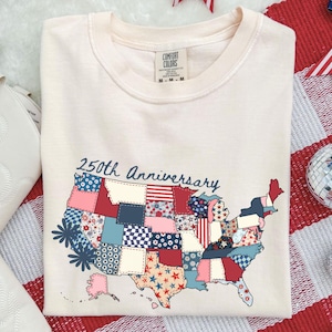 Pode incluir: Camiseta creme com um mapa de patchwork dos Estados Unidos, com padrões florais, de estrelas e listrados. O texto "250th Anniversary" está acima do mapa. A camisa é da Comfort Colors.