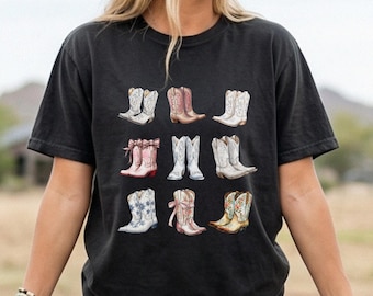 May include: A black short-sleeved t-shirt featuring a graphic print of nine different cowboy boots arranged in a grid. The boots are illustrated in various colors including white, pink, and tan, with details like bows, floral patterns, and stitching.