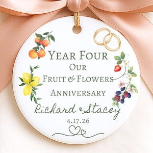 May include: A round white ceramic ornament with a peach ribbon, featuring watercolor illustrations of oranges, lemons, strawberries, and blackberries, two gold rings, and the text Year Four Our Fruit & Flowers Anniversary Richard & Stacey 4.17.26.