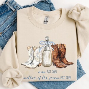 May include: Cream-colored sweatshirt with a watercolor design featuring white and brown cowboy boots, a champagne bottle with a blue bow, and the text "mom EST 2021" and "mother of the groom EST 2025". The sweatshirt is laid on top of blue jeans.