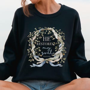 Puede incluir: Una sudadera negra de manga larga con un gr&aacute;fico central de una corona de flores entrelazada con cintas azul claro. El texto dentro de la corona dice HE RESTORES my soul PSALM 23:3 en una combinaci&oacute;n de fuentes serif y manuscritas.