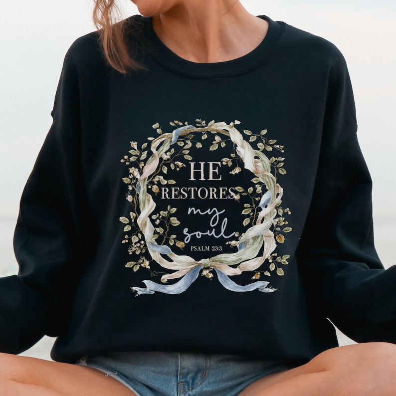 Puede incluir: Una sudadera negra de manga larga con un gr&aacute;fico central de una corona de flores entrelazada con cintas azul claro. El texto dentro de la corona dice HE RESTORES my soul PSALM 23:3 en una combinaci&oacute;n de fuentes serif y manuscritas.