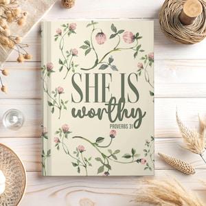 Op de afbeelding: Een hardcover notitieboek met een crèmekleurige kaft met de tekst "SHE IS worthy" in groen, met daaronder "PROVERBS 31". De kaft is versierd met aquarel illustraties van roze bloemen en groene bladeren.