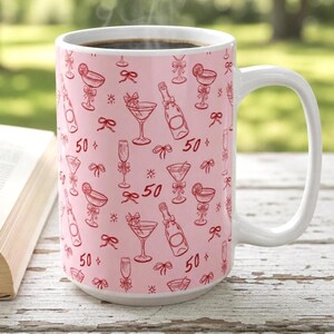 以下が含まれることがあります： コーヒーが入った白いセラミックマグ。ピンク色の背景に、赤いカクテルグラス、シャンパンボトル、リボン、数字の「50」が繰り返されるパターン。マグには大きな白いハンドルが付いています。