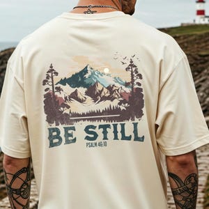 Op de afbeelding: Crèmekleurig T-shirt met een berglandschapontwerp op de achterkant. Het ontwerp bevat bomen, bergen en vogels, met de woorden "BE STILL" en "Psalm 46:10" eronder gedrukt. Het shirt heeft een neutrale kleur.