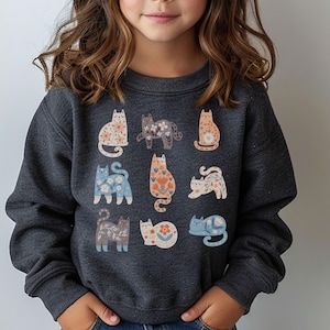 Puede incluir: Una sudadera de manga larga en color gris carbón oscuro con un estampado gráfico de nueve gatos estilizados en varias posturas. Cada gato está decorado con intrincados patrones florales y de hojas en tonos azul claro, naranja, crema y gris.