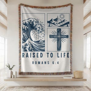 May include: A cream-coloured throw blanket with a navy blue graphic design. The design includes a wave, mountains, and a cross, with the text "RAISED TO LIFE ROMANS 6:4". The blanket has a fringed edge.