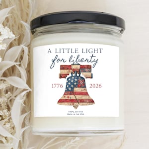 May include: A glass jar candle with a black lid. The label features the text "A LITTLE LIGHT for liberty" above an illustration of the Liberty Bell. The year 1776 and 2026 are printed below the bell. The candle is made with 100% soy wax.