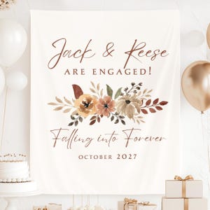 May include: A rectangular off-white fabric banner featuring the brown text Jack & Reese ARE ENGAGED! Falling into Forever OCTOBER 2027. The center displays a watercolor illustration of flowers and leaves in shades of orange, brown, and tan.