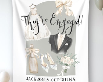 May include: A white fabric wall hanging featuring the text "They're Engaged!" and "JACKSON & CHRISTINA". Illustrations include a wedding dress, a tuxedo jacket, a champagne bottle with a bow, white heels, a floral bouquet, and a large cream bow.