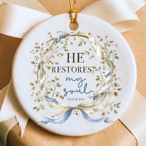 May include: A white ceramic Christmas ornament with the text "HE RESTORES my soul" and "PSALM 23:3". The ornament features a floral wreath design with ribbons in shades of blue, pink, and green, and a gold hanging loop.