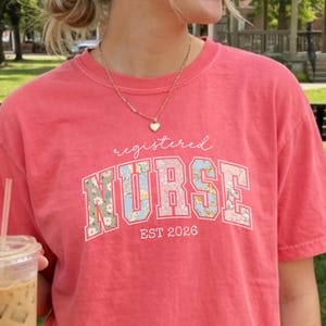 May include: A coral-colored t-shirt with the words "registered NURSE" in a floral design. The text "EST 2026" is below the word "NURSE". The shirt is a short-sleeved, crew neck style.