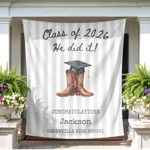 May include: A graduation banner with vertical gray and white stripes. The banner reads "Class of 2026 He did it!" and features a mortarboard hat atop a pair of brown cowboy boots. The banner also says "Congratulations Jackson Greenville High School."