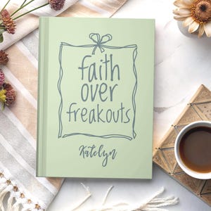 May include: A light green journal with the words "faith over freakouts" in a handwritten font, framed by a decorative border. The name "Katelyn" is written below the text. The journal is on a patterned surface.