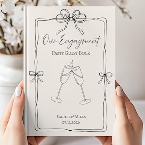 May include: A cream-coloured hardback guest book featuring a dark grey line-art border with bows, two clinking champagne flutes, and the text Our Engagment Party Guest Book, Rachel & Miles, and the date 07.12.2026 printed on the front cover.
