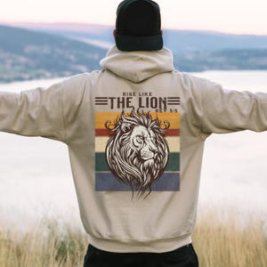 Op de afbeelding: Een beige hoodie met een zwarte pet. De achterkant van de hoodie heeft een leeuwenkopafbeelding met de tekst "RISE LIKE THE LION REV 5:5" boven een gestreepte achtergrond in retrostijl in tinten oranje, geel, groen en blauw.