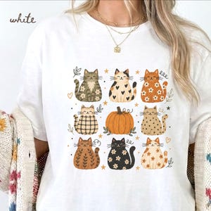 May include: A white t-shirt featuring a graphic print of eight stylized cats and a central orange pumpkin. The cats display various patterns including flowers, hearts, plaid, and stripes in shades of brown, black, and cream, accented by small stars.