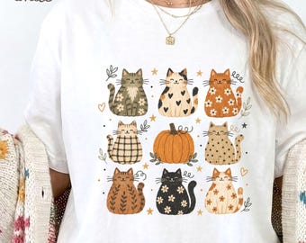 Puede incluir: Una camiseta blanca con un estampado gráfico de ocho gatos estilizados y una calabaza naranja central. Los gatos muestran varios patrones que incluyen flores, corazones, cuadros y rayas en tonos marrones, negros y crema, acentuados por pequeñas estrellas.