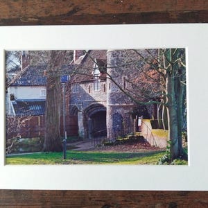 May include: A framed photograph of a stone archway and tower, possibly part of a historic building. The image is set against trees and grass, with a white mount and a wooden surface. The scene is bathed in sunlight.