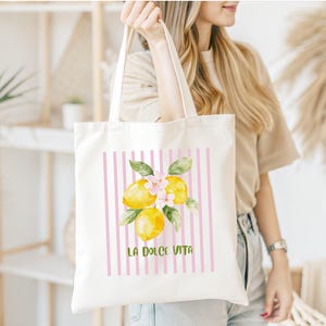 May include: A white tote bag featuring a watercolor illustration of lemons with pink flowers and green leaves. The design is set against a backdrop of pink vertical stripes, with the text "LA DOLCE VITA" in green.
