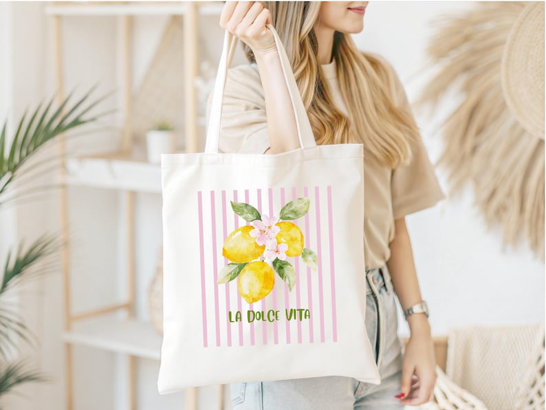 May include: A white tote bag featuring a watercolor illustration of lemons with pink flowers and green leaves. The design is set against a backdrop of pink vertical stripes, with the text "LA DOLCE VITA" in green.