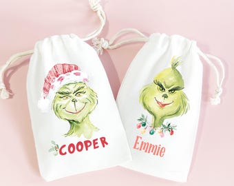 May include: Two white drawstring bags with Grinch character illustrations. One Grinch wears a Santa hat, the other has a garland of lights. The name "Cooper" is printed on one bag, and "Emmie" on the other, in red.