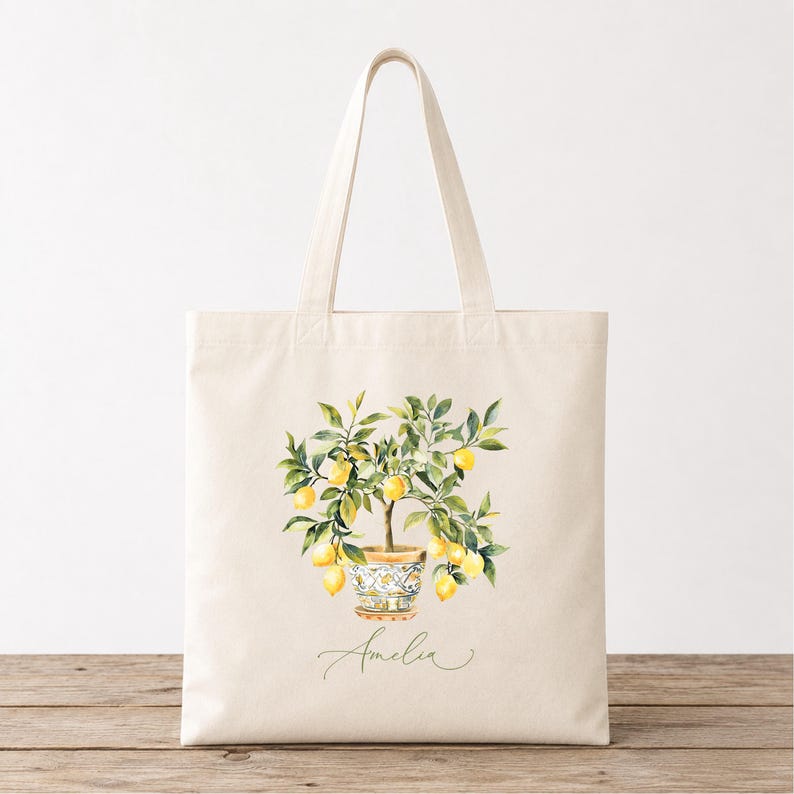 May include: Natural canvas tote bag with a watercolour illustration of a lemon tree in a patterned pot. The tree has yellow lemons and green leaves. The name "Amelia" is written in script below the tree.