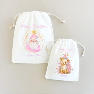 May include: Two white drawstring gift bags. The larger bag features a pink Santa Claus illustration, the text Merry Christmas, and the name Emmie Claire. The smaller bag shows a puppy in front of a pink Christmas tree, the text WE WOOF YOU A MERRY CHRISTMAS, and the name EMMIE.