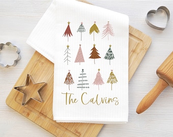 May include: White kitchen towel with a gold star and a red, pink, brown, and green Christmas tree design. The text "The Calvins" is printed in gold at the bottom of the towel.