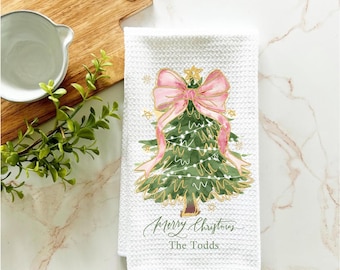 May include: White kitchen towel with a green and gold Christmas tree with a pink bow and lights. The text "Merry Christmas The Todds" is printed on the towel.