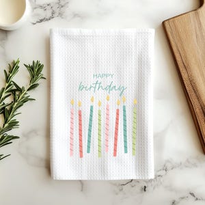 Puede incluir: Un paño de cocina blanco con tejido de gofre que presenta el texto Happy Birthday en letra cursiva de color verde azulado sobre una fila de nueve velas de cumpleaños delgadas y coloridas en tonos rosa, rojo, verde azulado y verde claro.